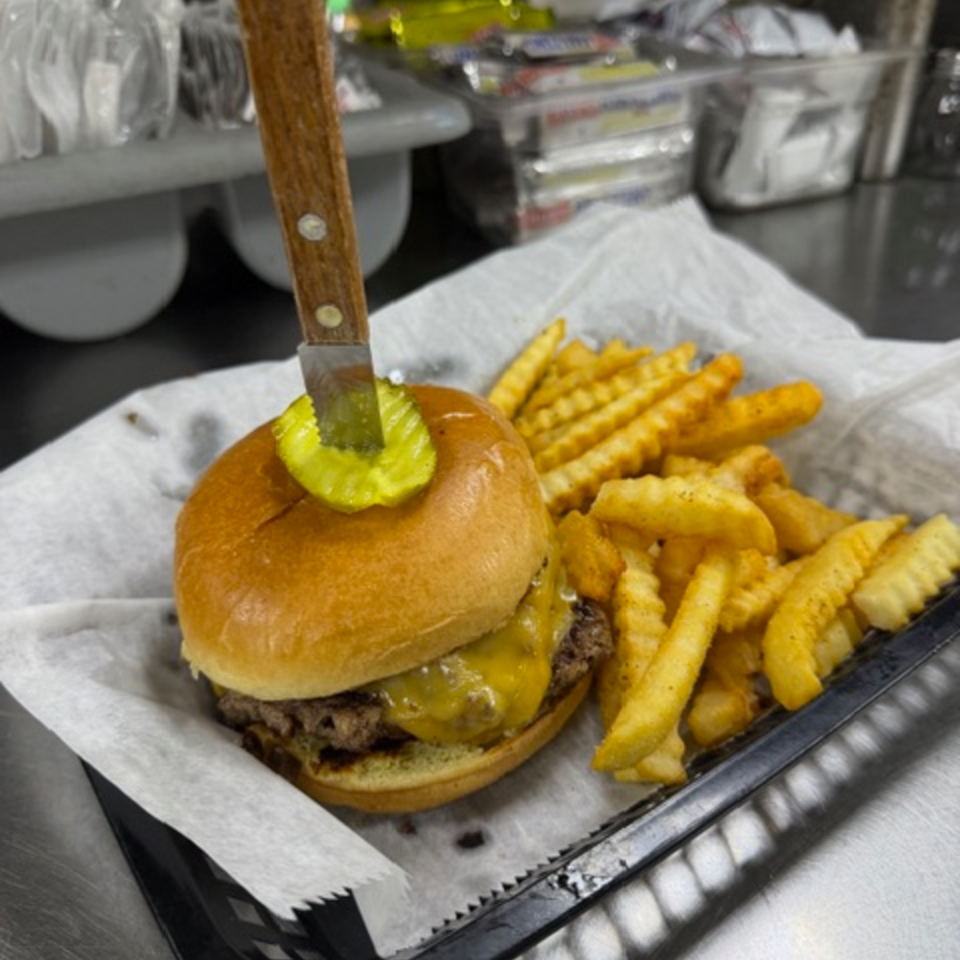 Cheeseburger with a pickle and knife on top, served with crinkle-cut fries in a basket.
