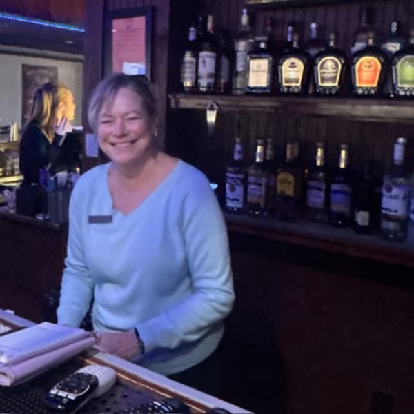 Smiling bartender in a light blue shirt behind a bar with liquor bottles on shelves.
