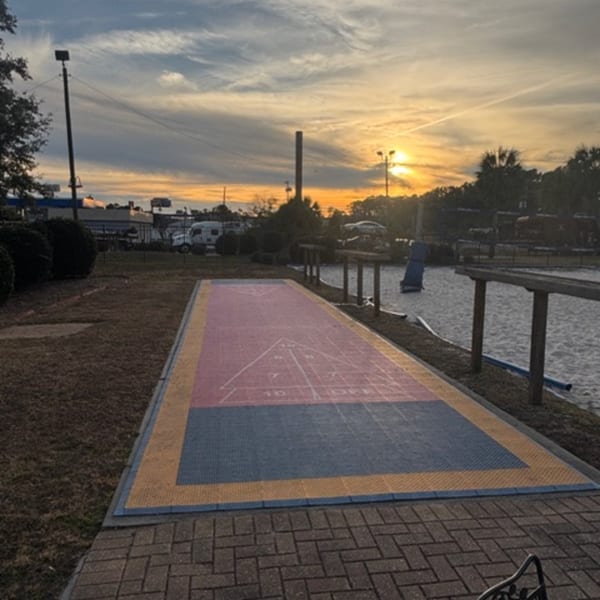 Outdoor shuffleboard court with pink, blue, and yellow sections under a sunset sky.