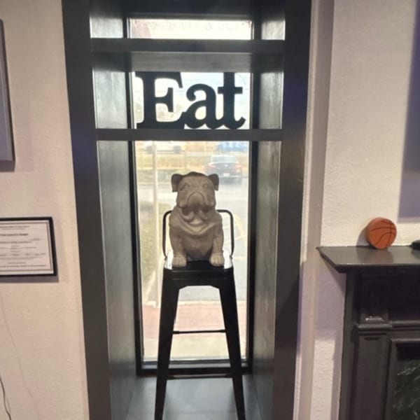 A bulldog statue on a bar stool in a window with an "Eat" sign above it.