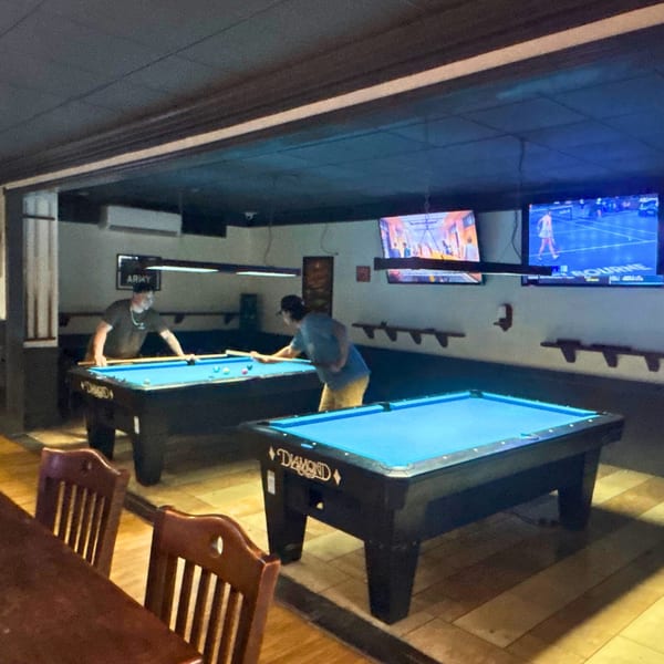 Two men play pool on blue-felt tables in a dimly lit room with TVs showing sports.