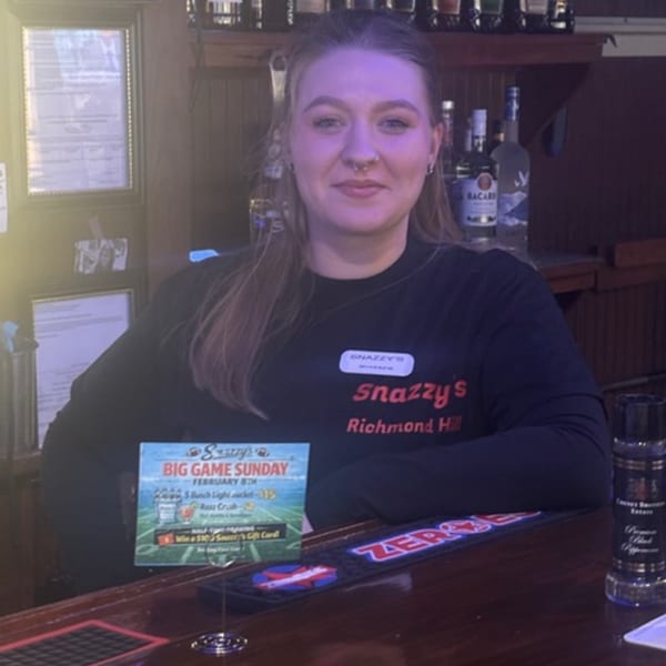 A Snazzy's Hot Wings employee in a black shirt stands behind a bar with a "Big Game Sunday" sign.