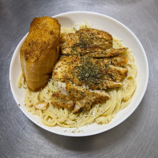 A white bowl of creamy fettuccine alfredo with seasoned chicken and a slice of garlic bread.