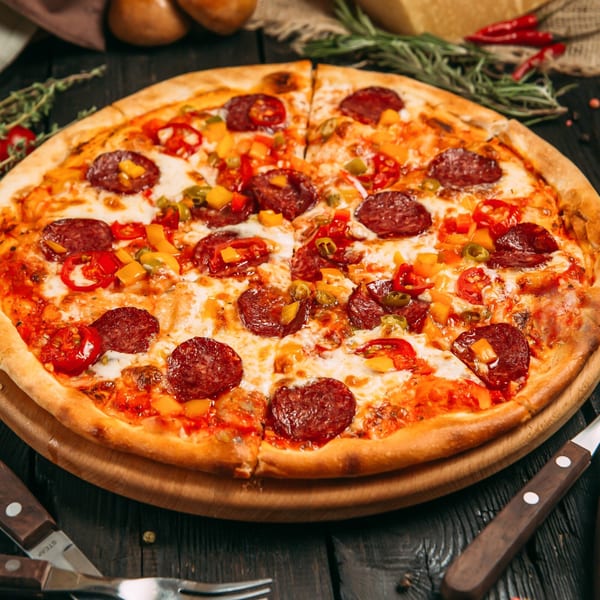 A whole pepperoni pizza with colorful bell peppers on a wooden board, with cutlery nearby.