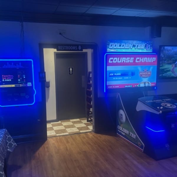 Arcade games with glowing blue lights, a restroom door, and checkered floor tiles at Snazzy's Hot Wings.
