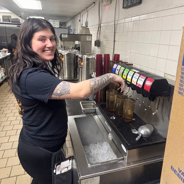 A smiling Snazzy's Hot Wings employee with tattooed arms fills drinks from a soda fountain.