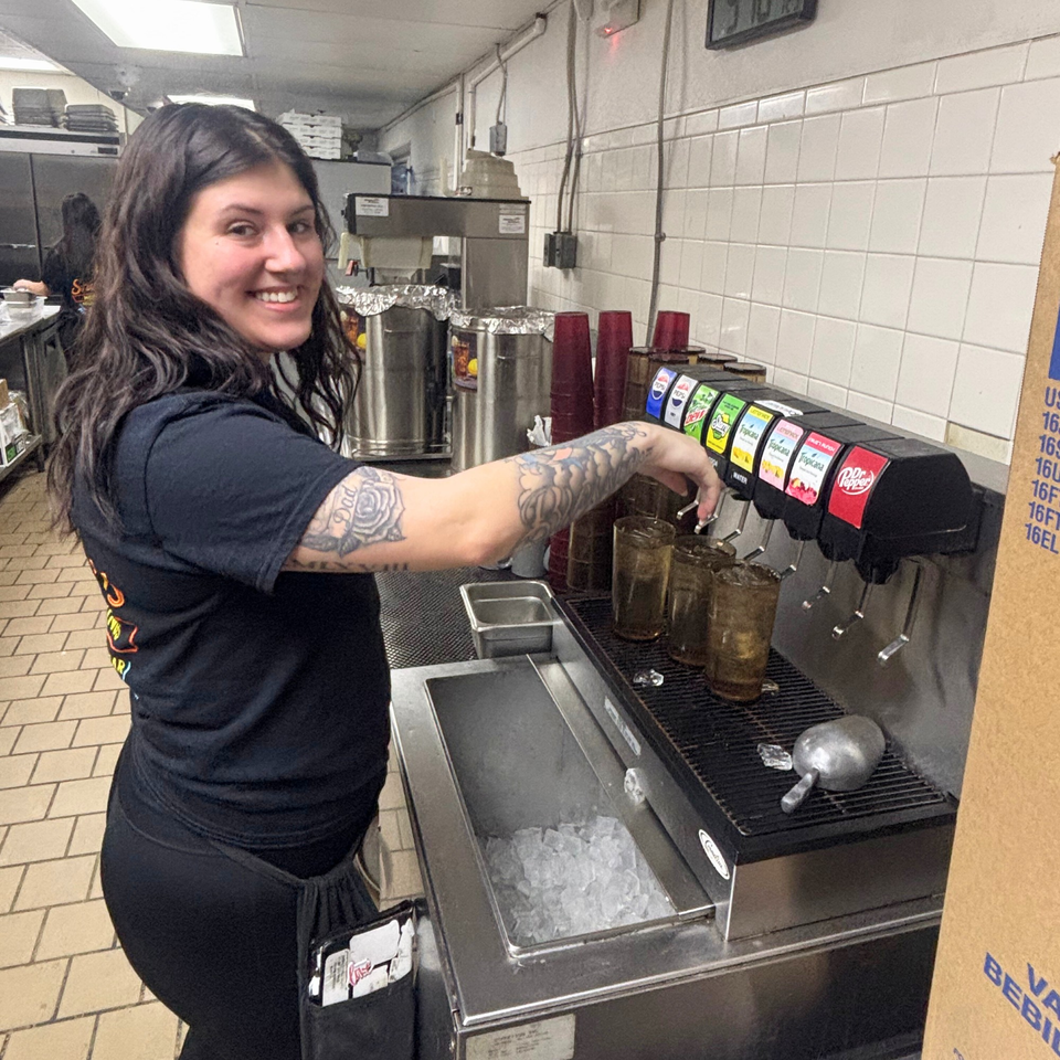 A smiling Snazzy's Hot Wings employee with tattooed arms fills drinks from a soda fountain.