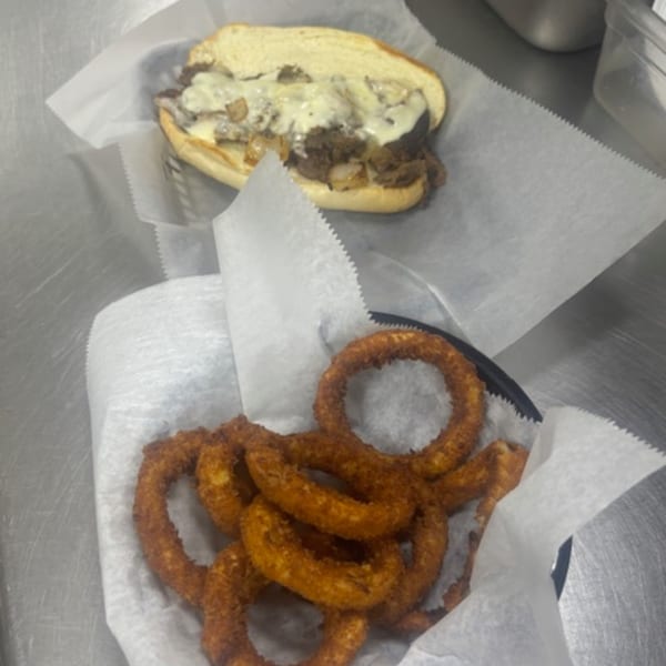 A Philly cheesesteak sandwich and a basket of crispy onion rings on a metal counter.