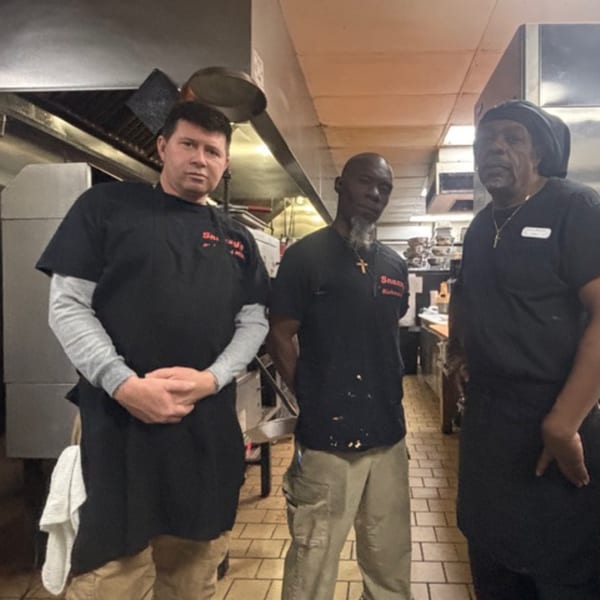 Three Snazzy's Hot Wings kitchen staff members in black shirts and aprons stand in the kitchen.