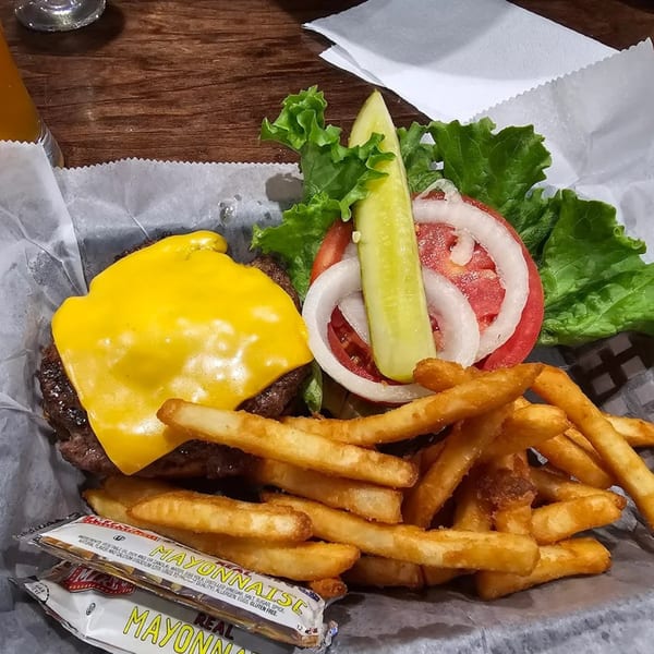 Cheeseburger with lettuce, tomato, onion, pickle, and fries in a basket with mayonnaise packets.